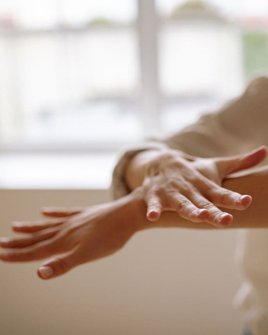 Des mains posées avec délicatesse sur un avant-bras, dans la lumière d'une fenêtre